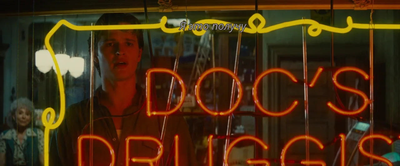 Making of: West Side Story (2021) movie still: medium — A man stands behind a glass window with a red neon sign reading Doc's Druggist. He is…