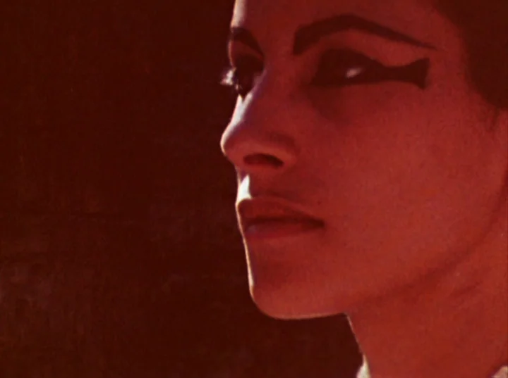Lucifer Rising (1972) movie still: closeup — A woman's face is shown in profile against a dark, featureless orange background. She wears dark…