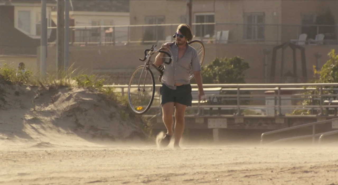 The Comedy (2012) movie still: fullbody — A sandy beach expanse sits in front of a distant wooden boardwalk and buildings. A man walks across the…