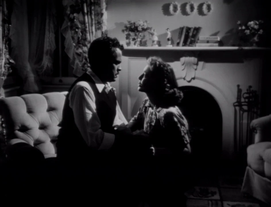 The Stranger (1946) movie still: medium — A dark living room with a fireplace and decorative objects on the mantle. A man and a woman sit together…