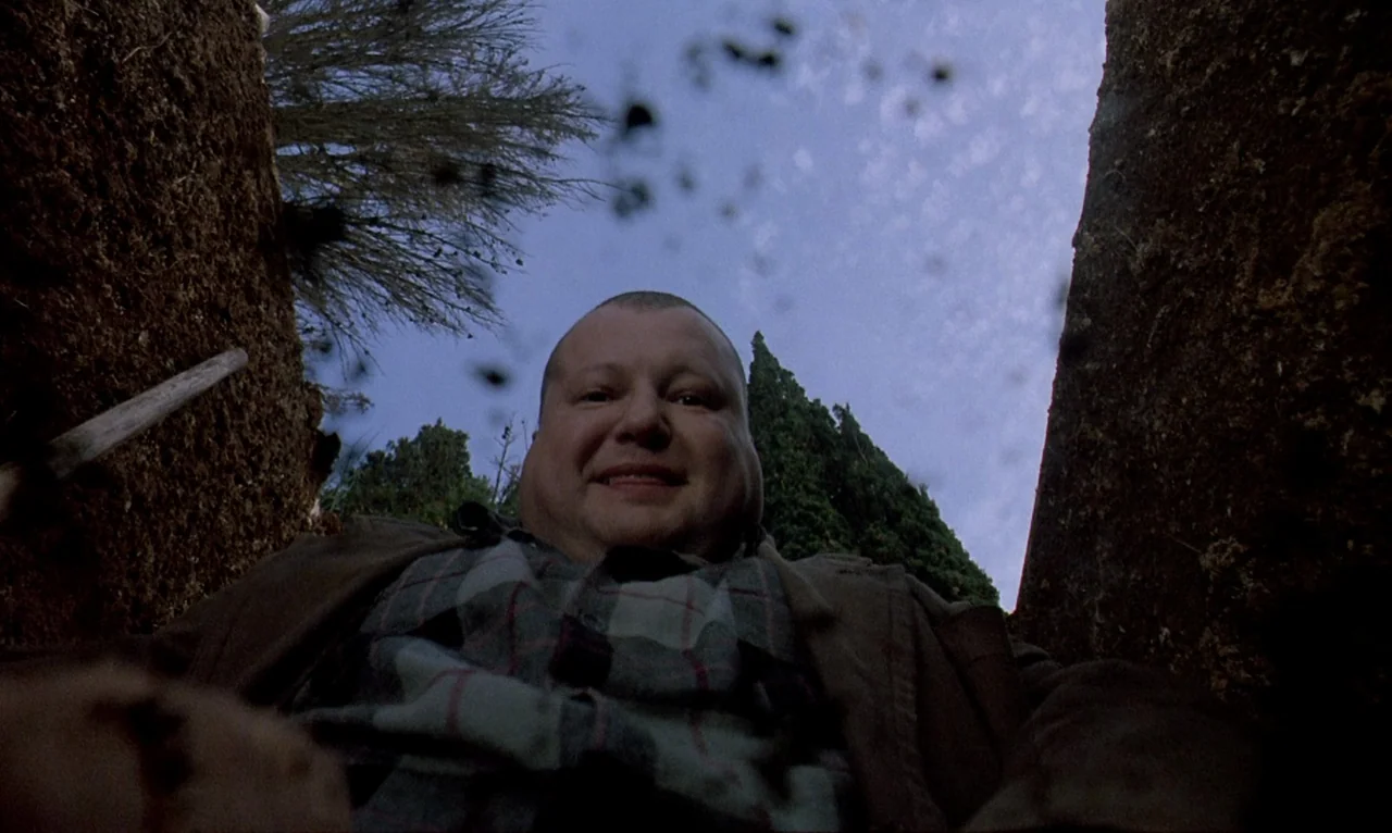 Dellamorte Dellamore (1994) movie still: medium — A man stands above an open dirt trench in a outdoor setting with trees. He gazes downward as loose…