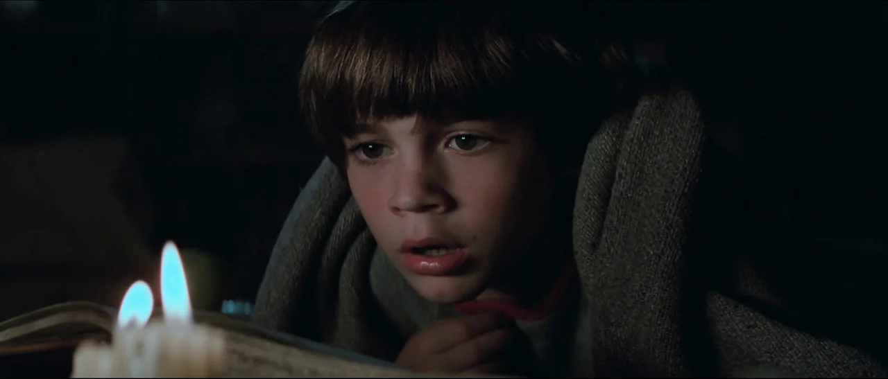 The NeverEnding Story (1984) movie still: closeup — A young boy reads an open book in a dark room under a blanket. The scene is illuminated only by…