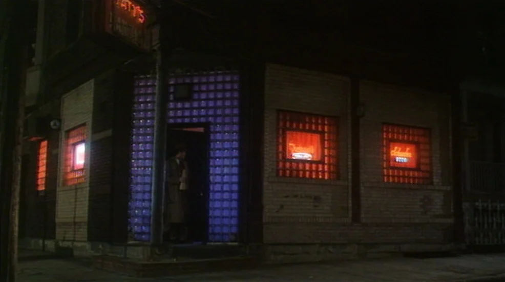 Union City (1980) movie still: wide — A nighttime street corner features a brick building with illuminated glass block windows. A man wearing a long…