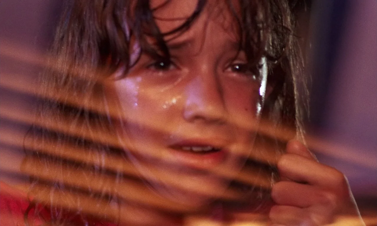 Demons 2 (1986) movie still: closeup — A dimly lit interior space is viewed through dark, horizontal window blinds. A child with wet hair looks…
