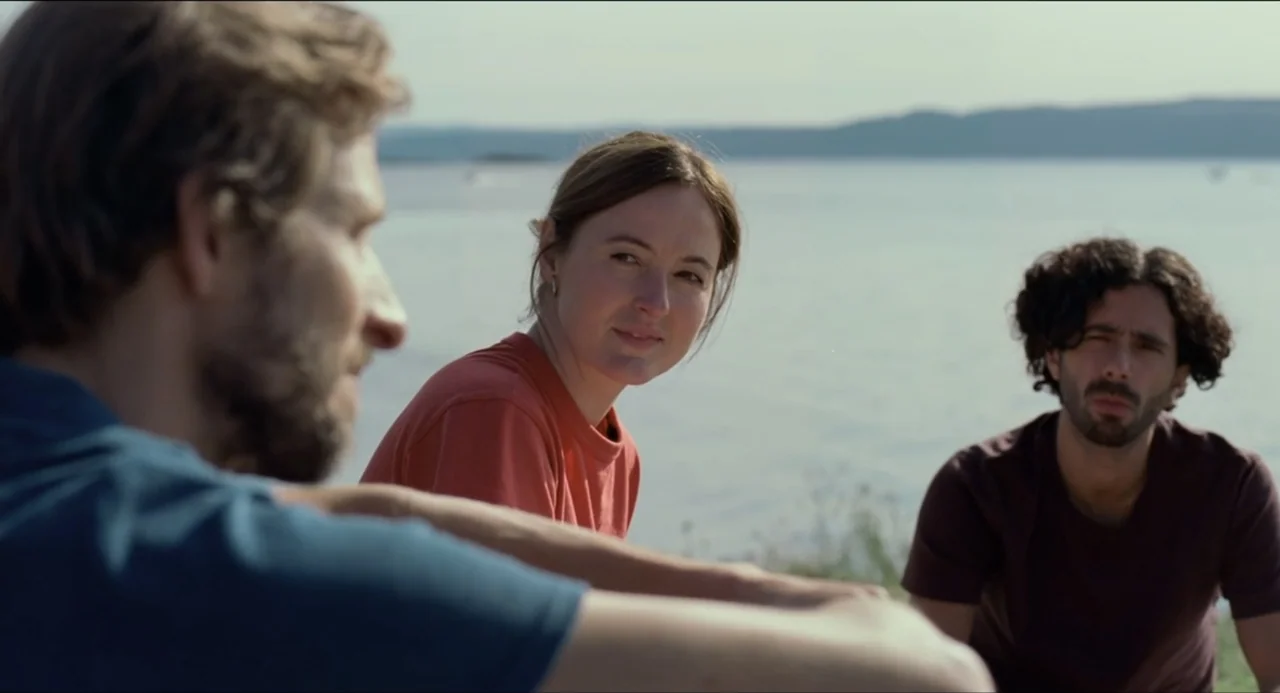 Sentimental Value (2025) movie still: medium — Three people sit on a grassy bank overlooking a calm lake in daylight. They are positioned in a…