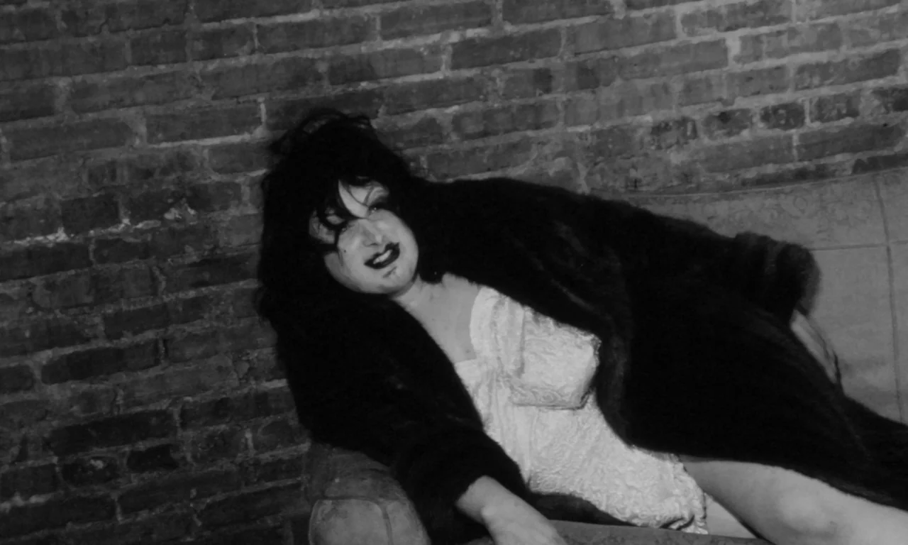Multiple Maniacs (1970) movie still: medium — An interior space features a rough exposed brick wall behind a plush sofa. A person with theatrical…
