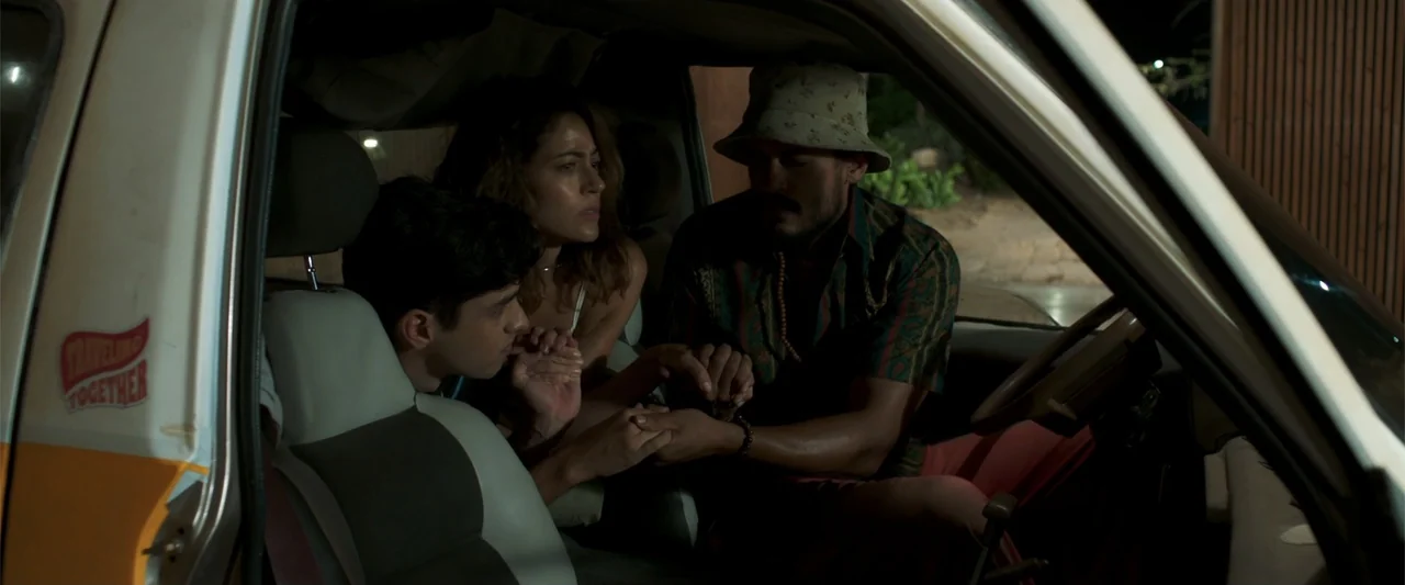 Férias Trocadas (2024) movie still: medium — The interior of a parked van at night with three individuals seated in the front row. They hold hands…