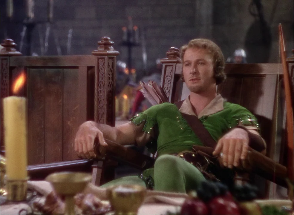 The Adventures of Robin Hood (1938) movie still: medium — A stone-walled interior space with wooden furniture and a dining table filled with food. A…