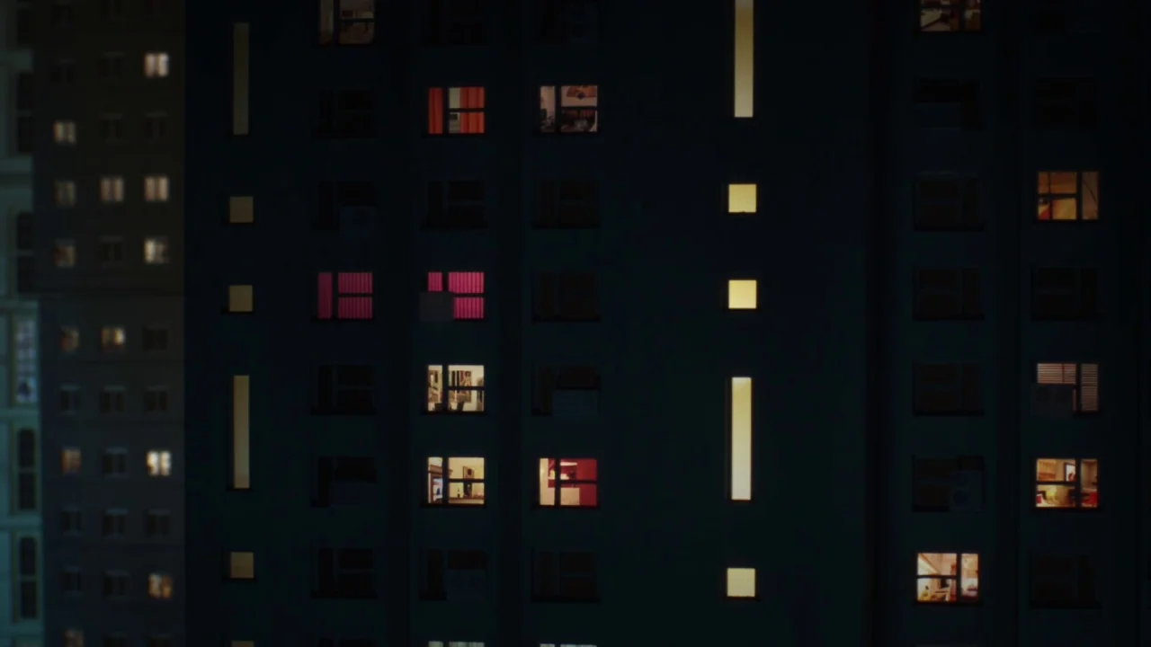 Piercing (2018) movie still: establishing — A high-rise residential building facade at night features dark walls and rectangular windows. Many of…