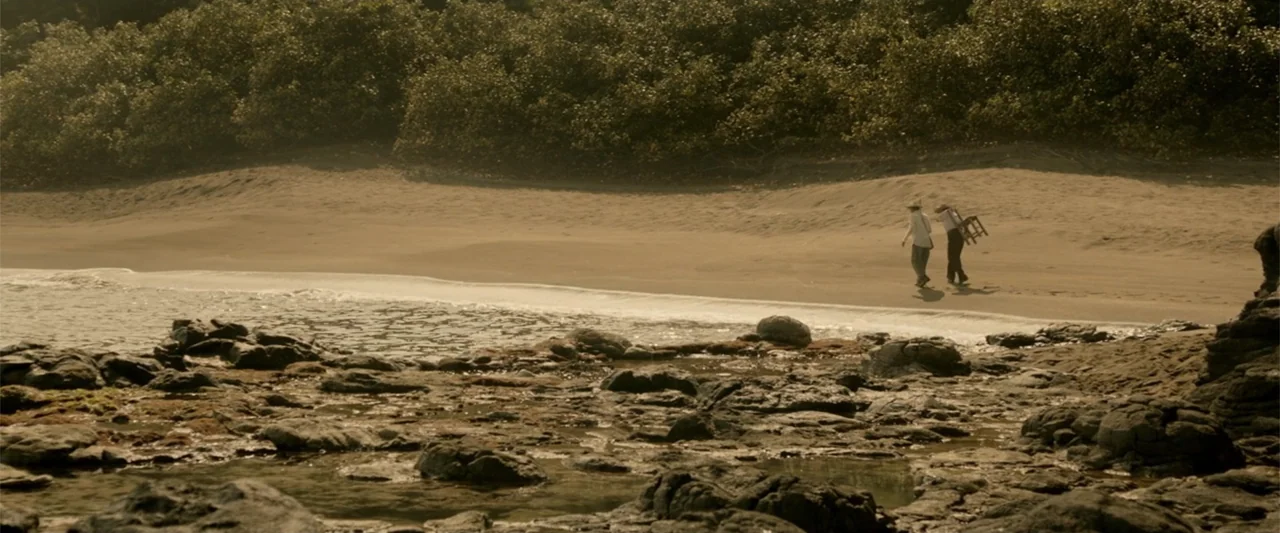 I Dream in Another Language (2017) movie still: wide — A remote sandy beach borders a dense line of dark green tropical vegetation. Two figures walk…