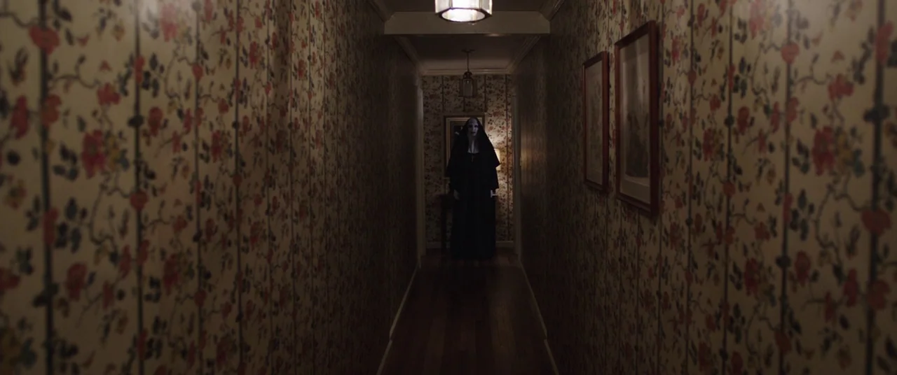 The Nun (2018) movie still: wide — A long, narrow interior corridor features walls covered in vintage floral wallpaper. A figure in a black nun…