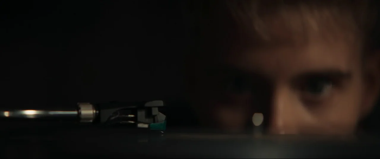 Evil Dead Rise (2023) movie still: closeup — A dark space features a record player with a tonearm resting on a disc. A man's out-of-focus face is…