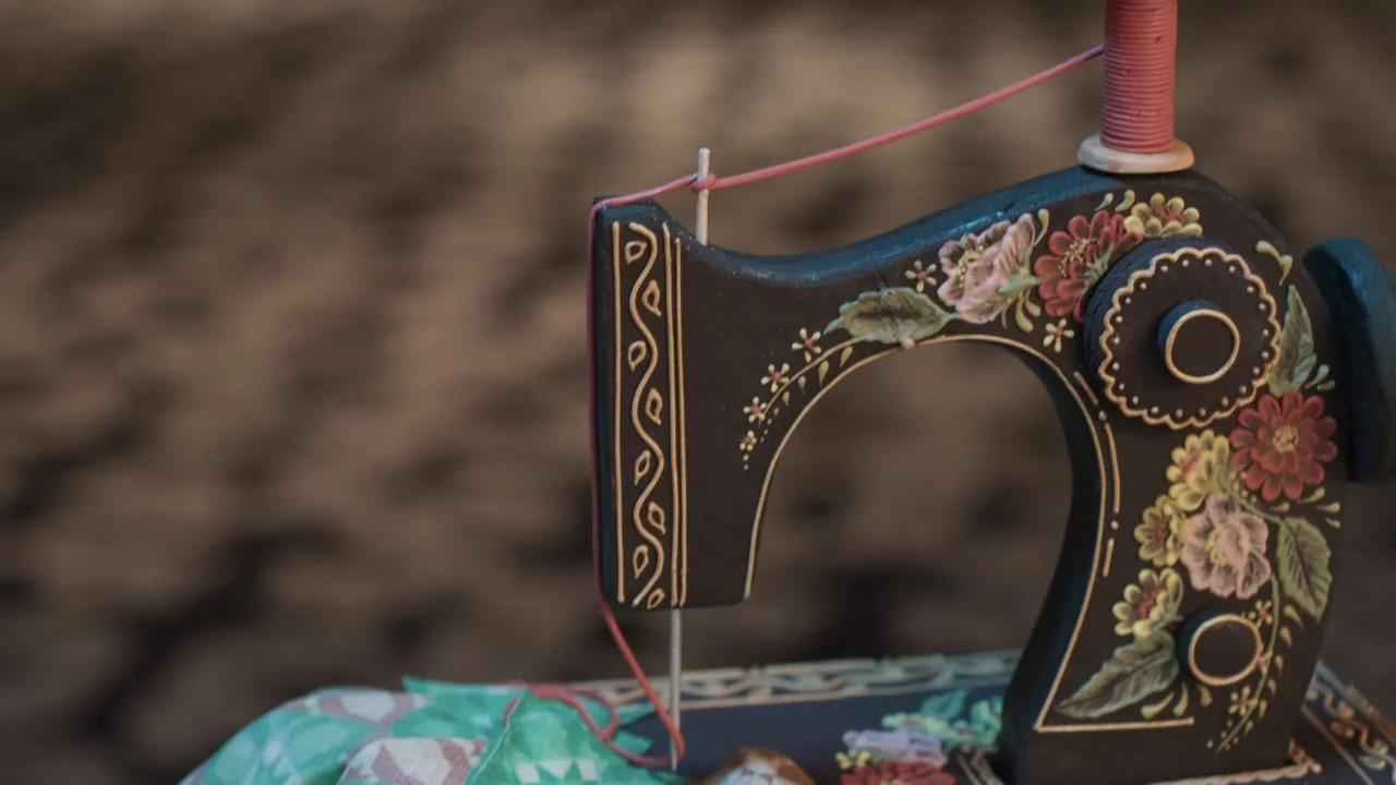 A Gingerbread Christmas (2022) movie still: closeup — A close view of a dark wooden sewing machine with hand-painted floral details. A spool of pink…
