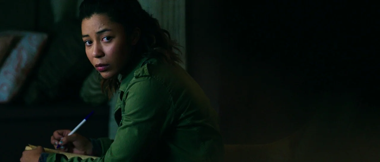 The Purge: Anarchy (2014) movie still: medium — A dimly lit room contains a woman seated and writing in a notebook. The surrounding area is obscured…
