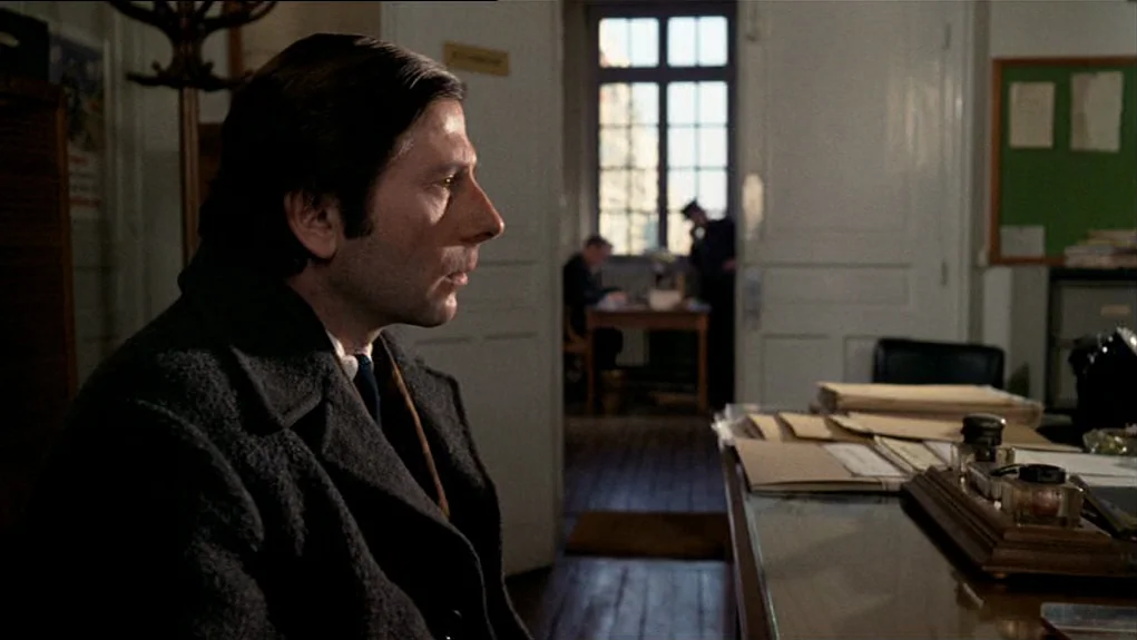 The Tenant (1976) movie still: medium — A quiet administrative room contains wooden desks, stacks of paper, and an inkwell set. A man in a heavy…