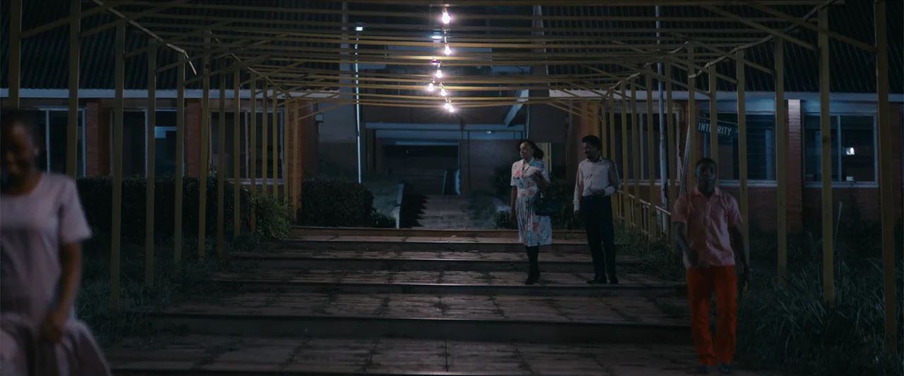 Ms. Kanyin (2025) movie still: wide — An exterior covered walkway at night features a series of overhead light bulbs. Several people in…