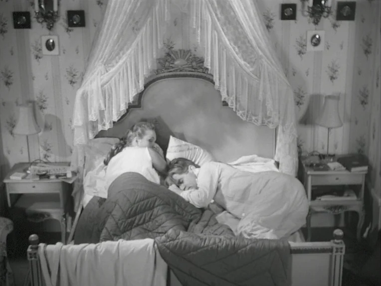 A Lesson In Love (1954) movie still: medium — A vintage bedroom features floral patterned wallpaper and a decorative bed with a canopy. A man and…