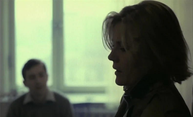 No End (1985) movie still: medium — A spare interior space contains a large window with white frames in the background. A woman in the foreground is…
