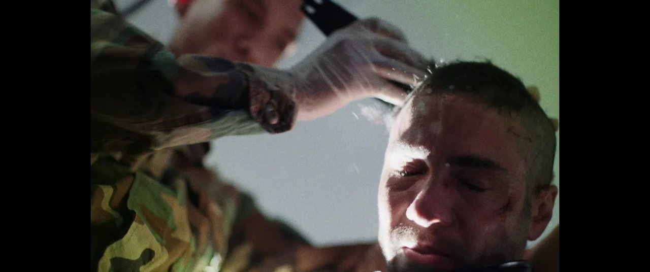 The Mauritanian (2021) movie still: closeup — An indoor space with a man receiving a military-style buzz cut. A person wearing a camouflage uniform…