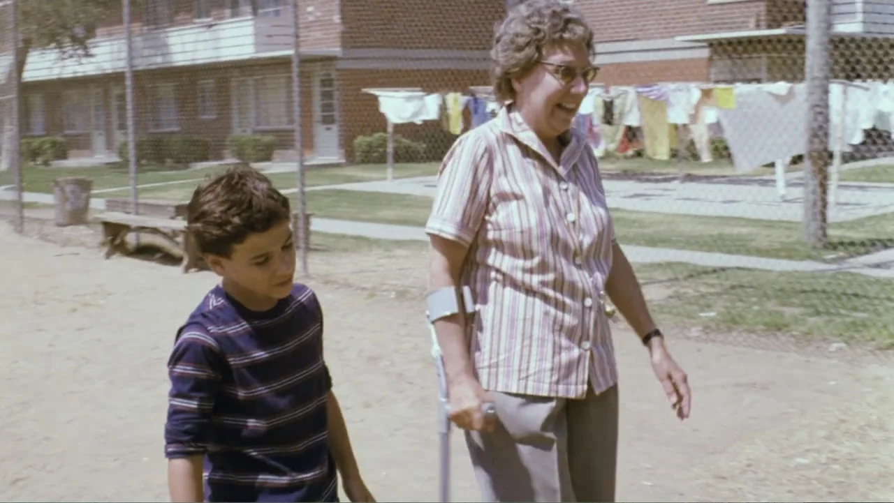 Aunt Mary (1979) movie still: fullbody — An outdoor playground features a chain-link fence and laundry hanging in the background. A woman with a…