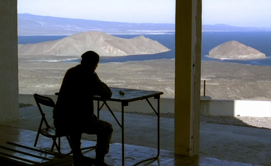 Beau Travail (1999) movie still: wide — A concrete patio overlooks a large body of water with distant rocky islands. A man sits at a simple folding…