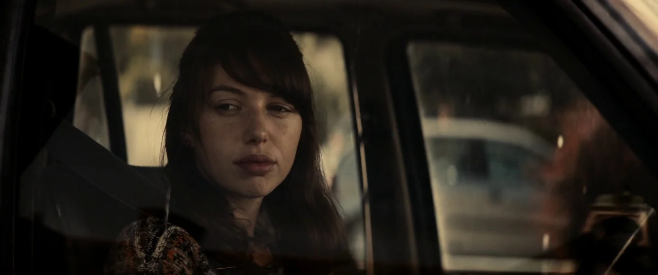 The Hole in the Ground (2019) movie still: medium — A woman sits inside a car during the day with blurred urban surroundings outside. She looks out…