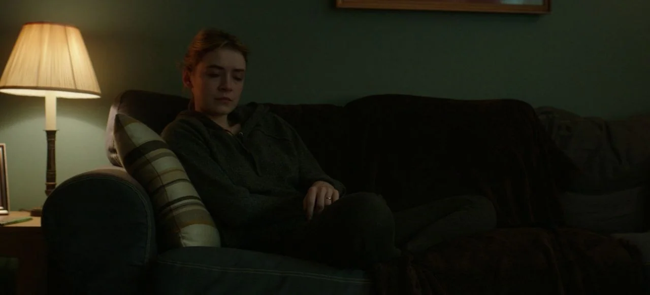 A Good Woman Is Hard To Find (2019) movie still: medium — A dark, dimly lit room contains a person sitting on a comfortable couch. A table lamp with…