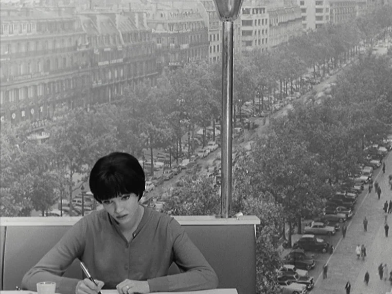 Vivre Sa Vie (1962) movie still: medium — A woman sits at a table in a cafe with a large window view. Outside, a busy Parisian street with trees and…