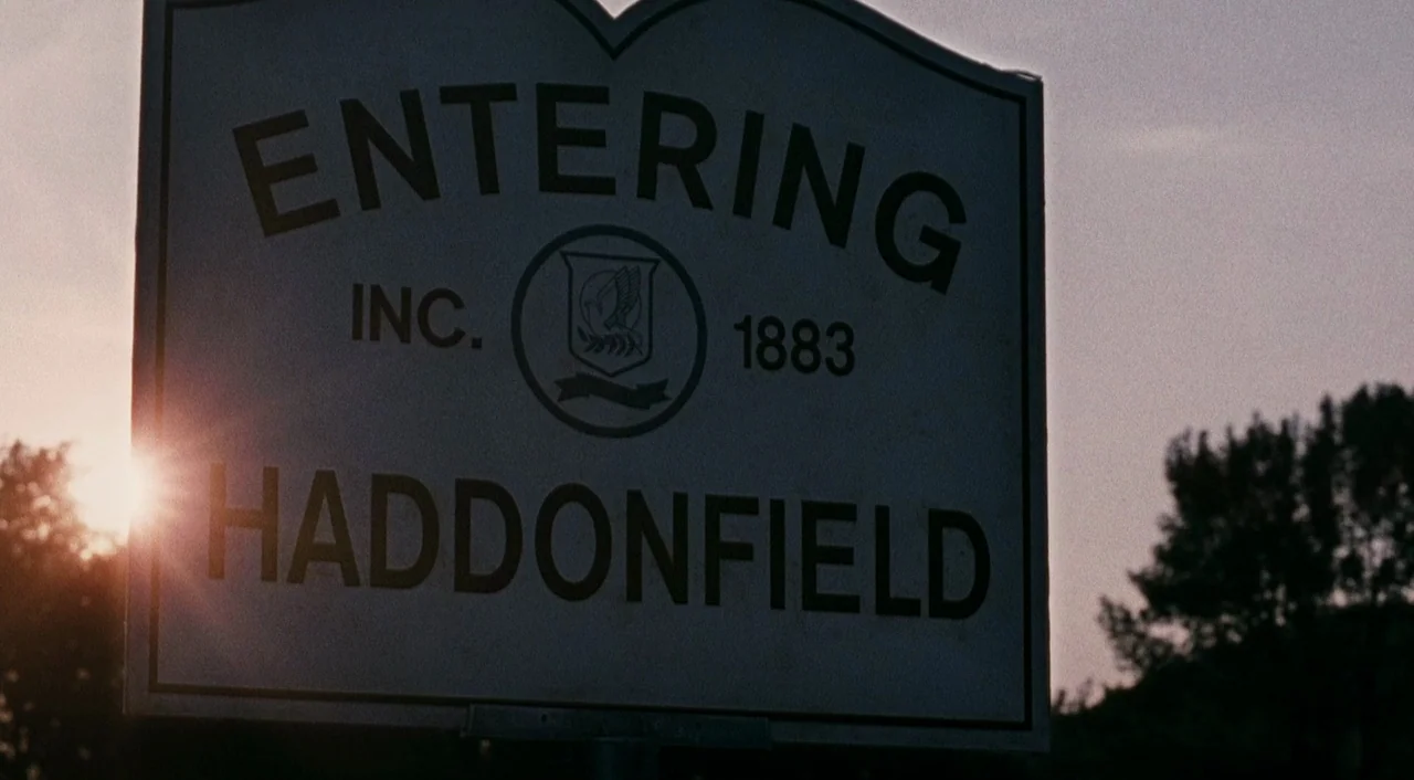 Halloween 2 (2009) movie still: closeup — A metal town sign on a post stands against a hazy sunset horizon. The sign displays the text Entering…