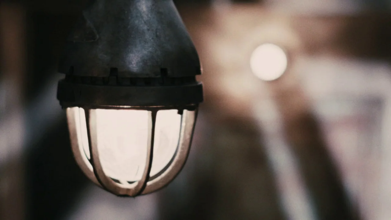 Saw VI movie still: closeup — An industrial cage light hangs in the foreground within a darkened room. The background is out of focus with soft…