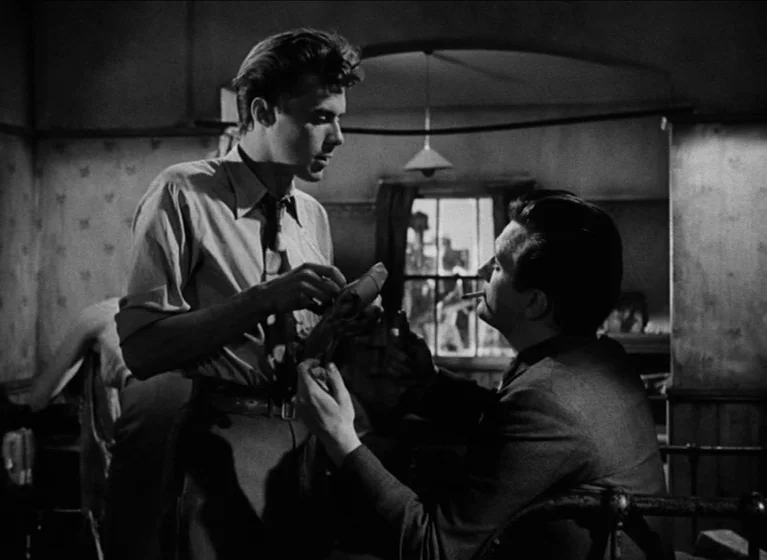 The Blue Lamp (1950) movie still: medium — A sparsely furnished room features textured walls and a small window with visible exterior detail. A man…