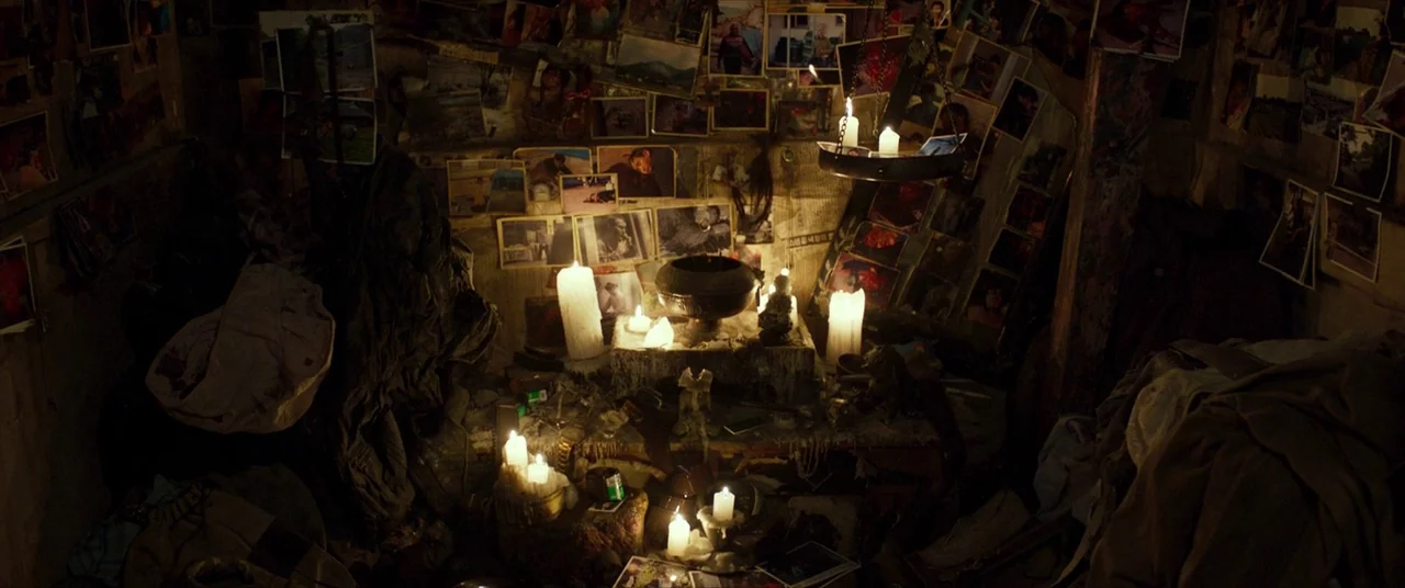 The Wailing (2016) movie still: wide — A dark, cluttered room is covered in numerous pinned photographs and lit by candles. A central altar holds a…