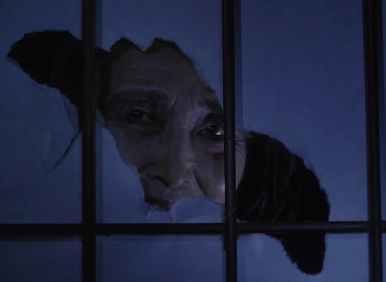 Pastoral To Die In The Country (1974) movie still: closeup — An elderly person gazes through a jagged hole torn in a paper wall surface. Black metal…