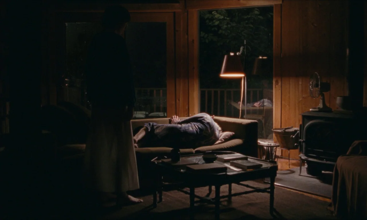 Janet Planet (2023) movie still: wide — A dark, wood-paneled living room at night features a floor-to-ceiling glass window wall. A man rests on a…