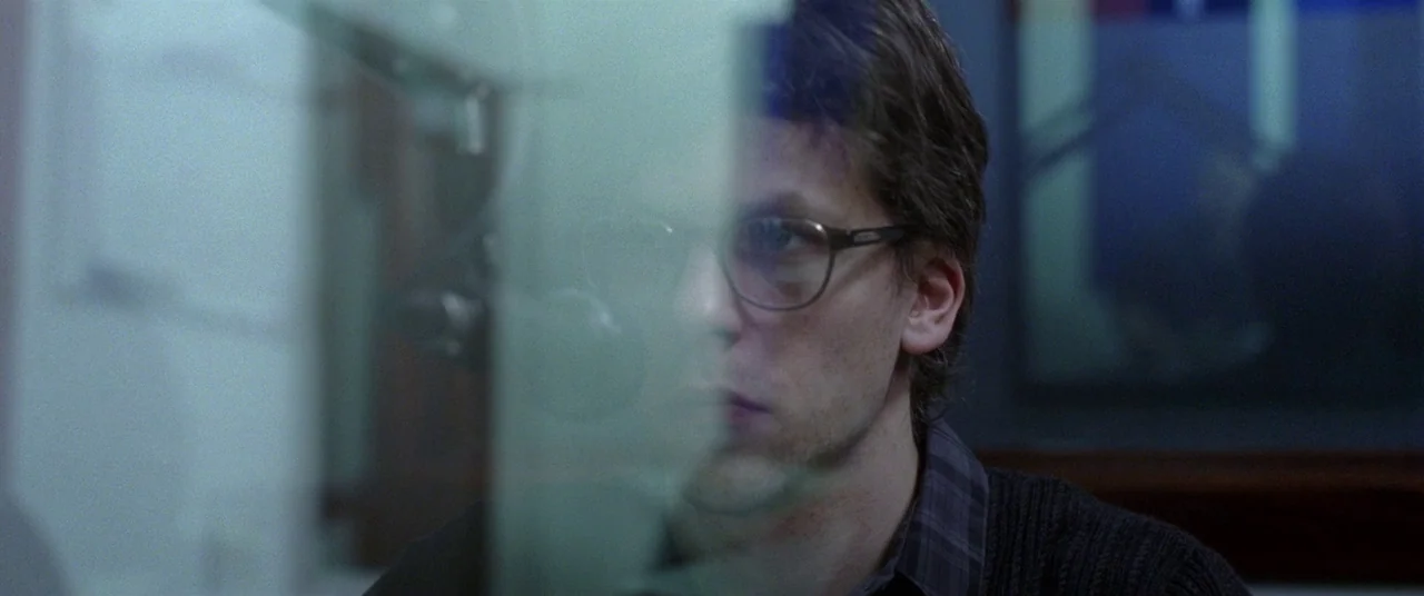 The End of the Tour (2015) movie still: closeup — A dimly lit room features a person positioned behind a reflective glass panel. The subject, a…