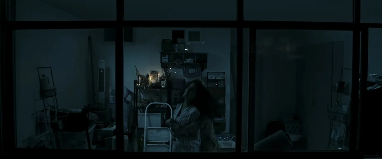 Evil Eye (2020) movie still: medium — A dimly lit room viewed through a large dark-framed window grid at night. A woman in pajamas stands alone…