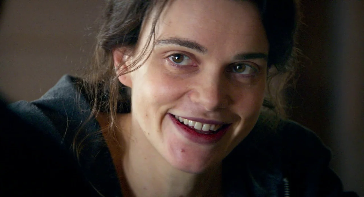 Anatomy of a Fall (2023) movie still: closeup — An interior space with a dark background and ambient light. A woman in a black jacket smiles at the…