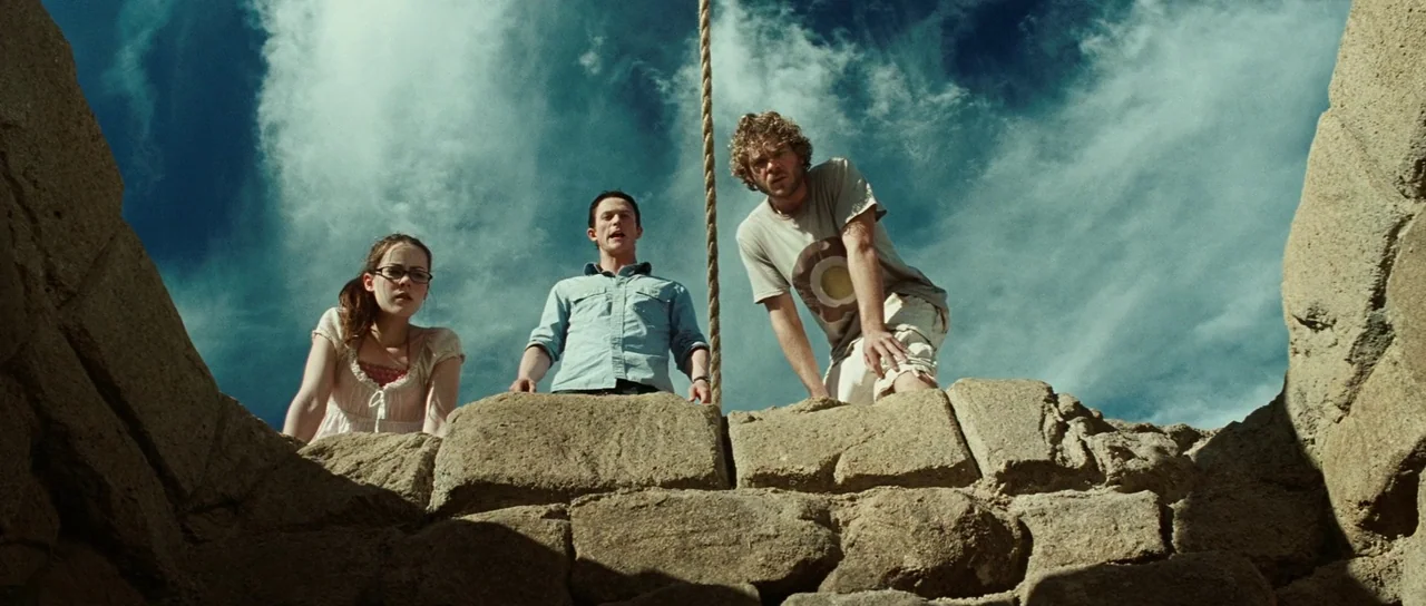 The Ruins (2008) movie still: medium — Three people stand at the edge of a deep stone shaft under a bright sky. They look down into the dark abyss…
