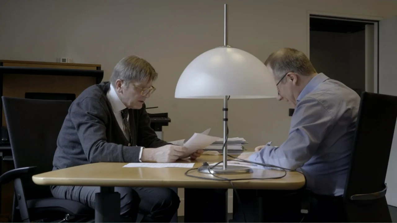 Brexit: Behind Closed Doors (2019) movie still: medium — A plain corporate office features a light wood table and a modern dome lamp. Two men in…