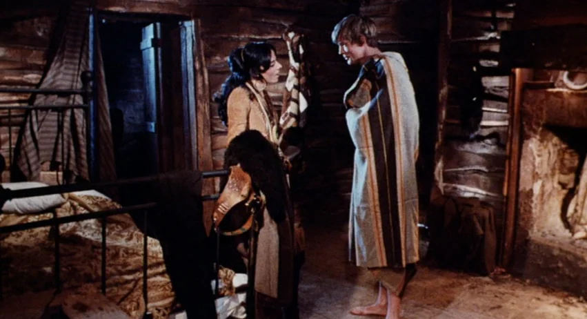 Roy Colt and Winchester Jack (1970) movie still: medium — A rustic log cabin interior features rough-hewn timber walls and a stone fireplace. A man…