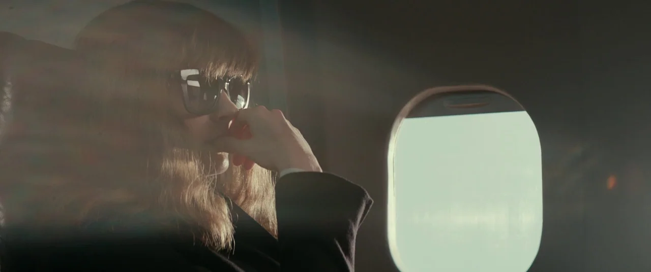 Colossal (2016) movie still: closeup — A close-up interior shot inside an airplane cabin showing a window. A woman with blonde hair and sunglasses…
