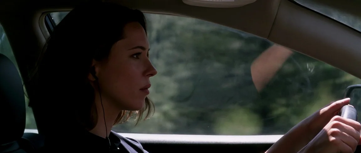Transcendence (2014) movie still: medium — The interior of a moving car shows a woman driving along a road. The woman wears small earphones while…