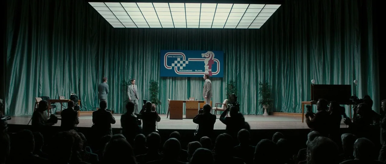 Pawn Sacrifice (2014) movie still: wide — A wide interior view of a chess tournament stage with teal curtains and banners. Two men stand by a table…