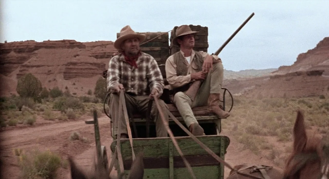 Ride In The Whirlwind (1966) movie still: fullbody — Two men ride on a wooden wagon through a vast arid desert landscape. The men hold leather reins…