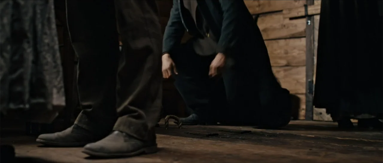 Out of Liberty (2019) movie still: medium — A dark wooden room with rough-hewn plank walls and floorboards. One man in an overcoat stands while…