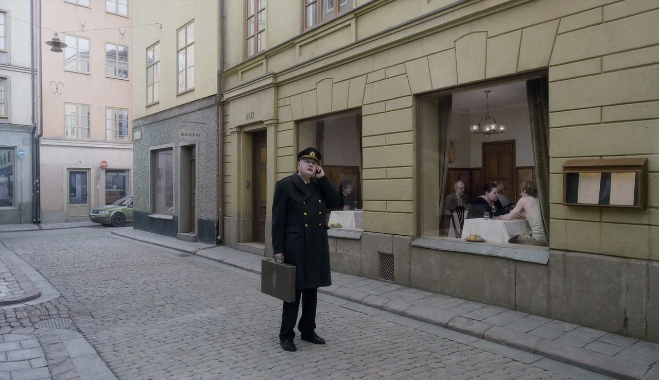 A Pigeon Sat on a Branch Reflecting on Existence (2014) movie still: fullbody — A quiet cobblestone street features a pale yellow building with a…