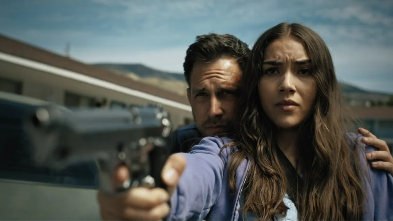 Bullet Proof (2022) movie still: medium — A sunny motel parking lot with a low building visible in the background. A woman in a blue hoodie points a…