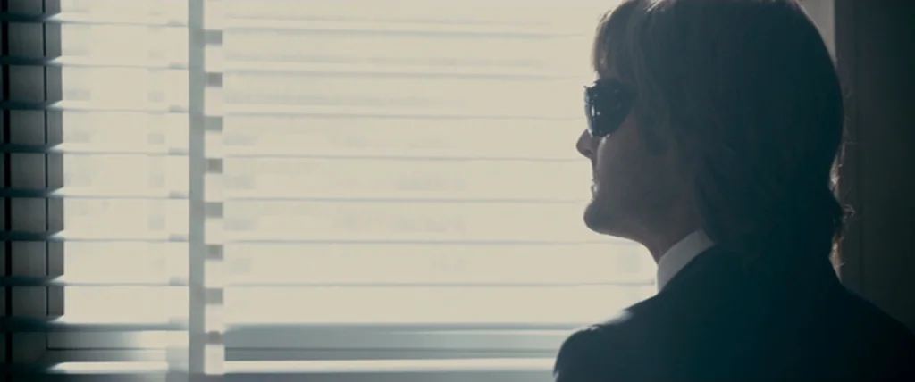 MacGruber (2010) movie still: medium — A dark, professional space with closed horizontal blinds filtering bright light from a window. A man in a…