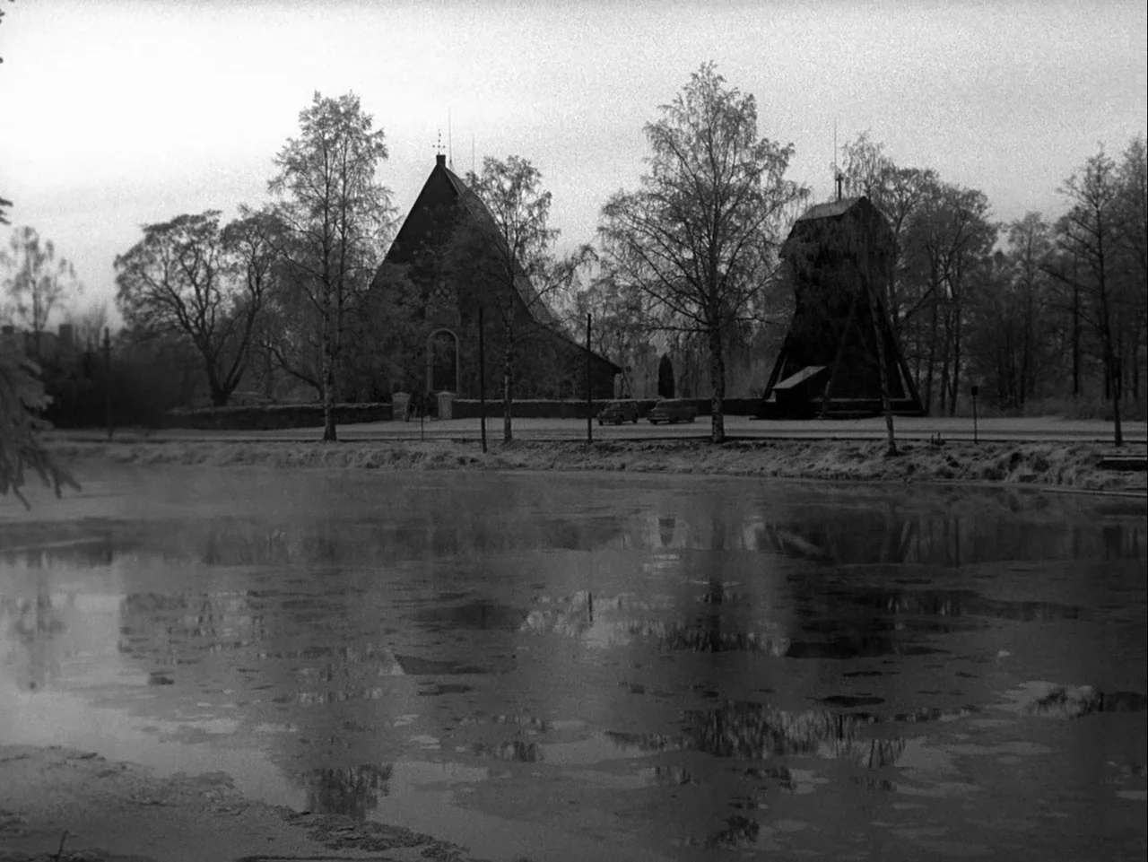 Winter Light (1963) movie still: establishing — A dark church building stands behind a calm, partially frozen pond with skeletal trees. An wooden…