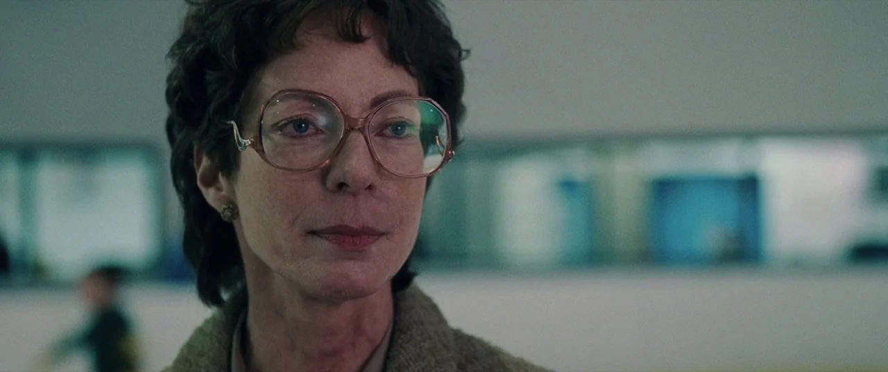 I, Tonya (2017) movie still: closeup — An indoor ice arena with blurred glass partitions and blue-tinted rink markings behind. A woman with glasses…